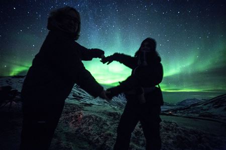 Two people forming a heart with their hands under the Northern Lights in a snowy mountain landscape at night.