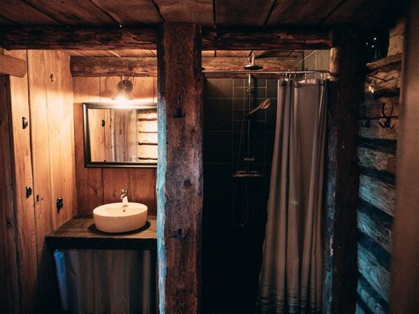 Litet porslinshandfat med spegel och dusch med grått kakel och synliga timmerväggar och massiva trästolpar.  