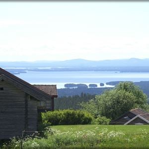 View over the lake with a gray timber cottage and a green meadow in the foreground. 