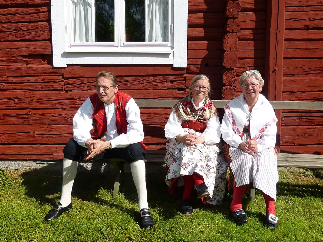 A man and two women sitting in front of a red house dressed in traditional clothes.
