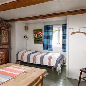 Rum med gammalt skåp med dalmålningar, en enkelsäng, en pinnstol och ett matbord. 