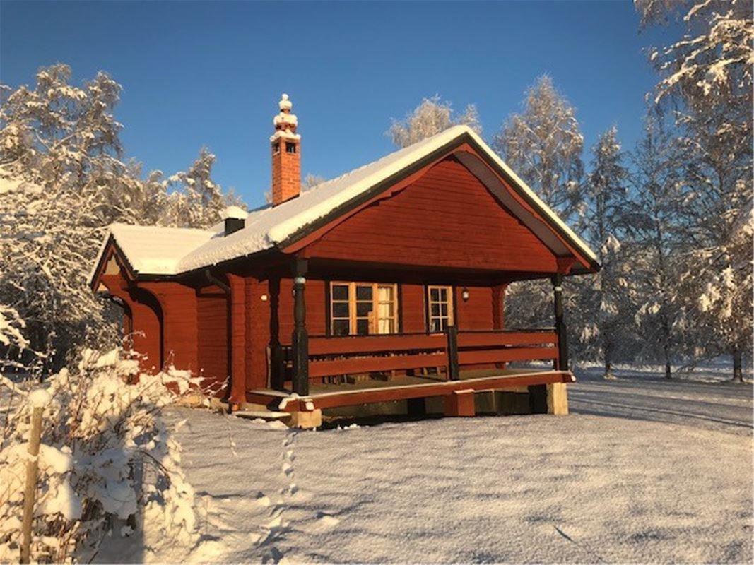 Röd stuga med veranda under tak och snö på taket och träden. 