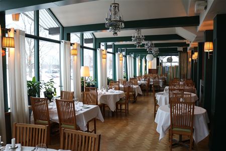 Tables with white cloths and wooden chairs in a restaurant with large windows.