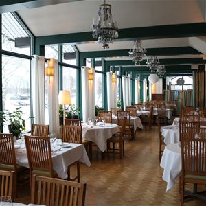 Tables with white cloths and wooden chairs in a restaurant with large windows.