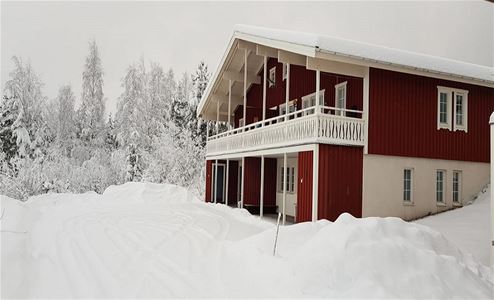 Rött suterränghus med vita fönsterfoder och balkong på övervåningen och snöklädda träd i bakgrunden.