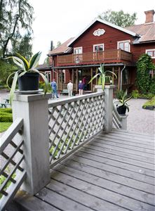En vit liten bro, en grusgång, tre personer som tittar på en töd träbyggnad i två våningar med veranda och balkong ovanpå verandan.