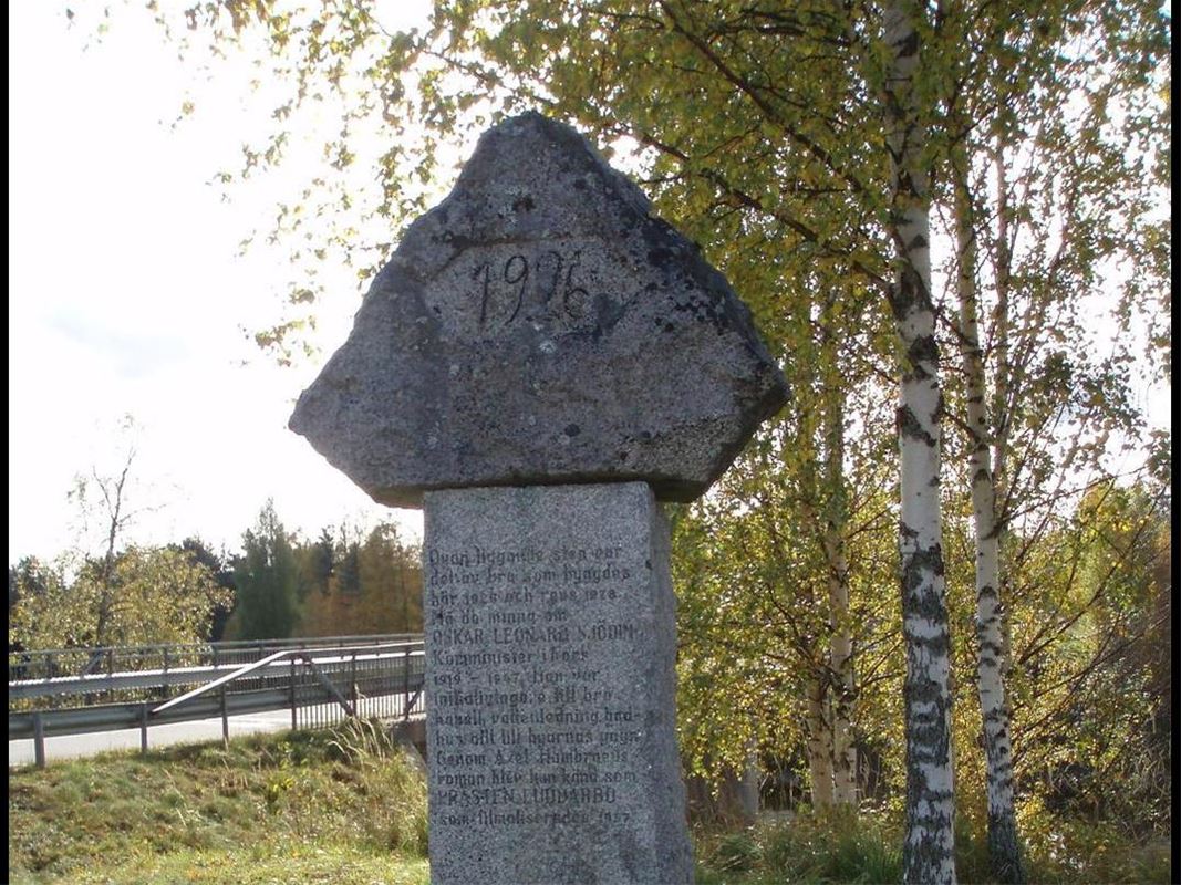 The memorial stone with birch in greenery and bridge behind.