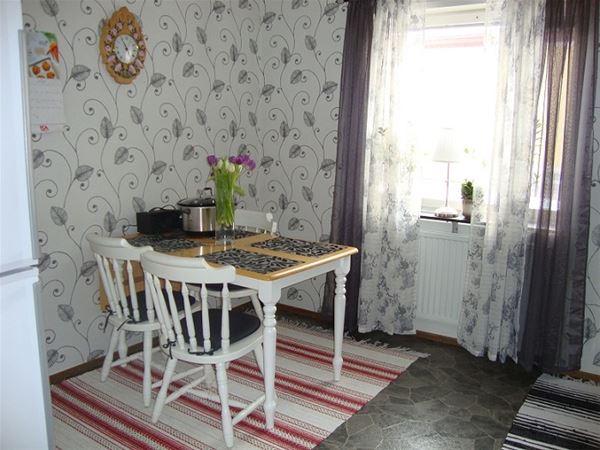 Matbord med 3 vita stolar i ett kök med en tapet med grått blom-mönster och gråa gardiner.  