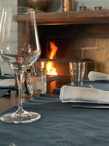 A glass in the foreground on a set table and a fire is burning in the fireplace.
