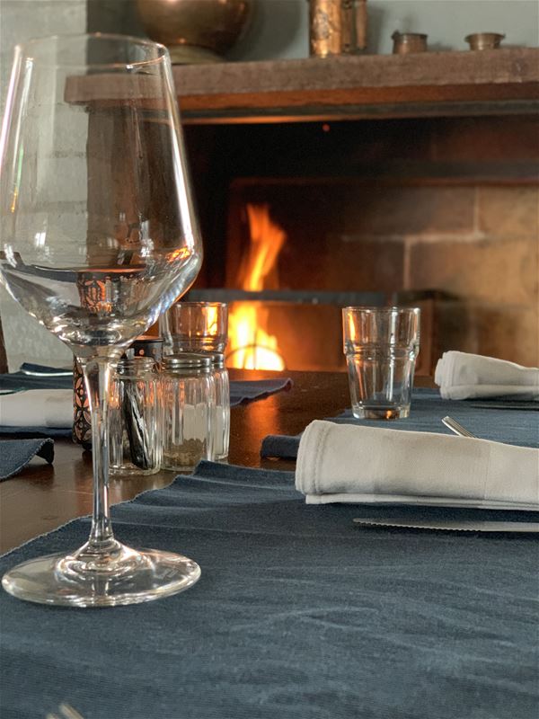 A glass in the foreground on a set table and a fire is burning in the fireplace.