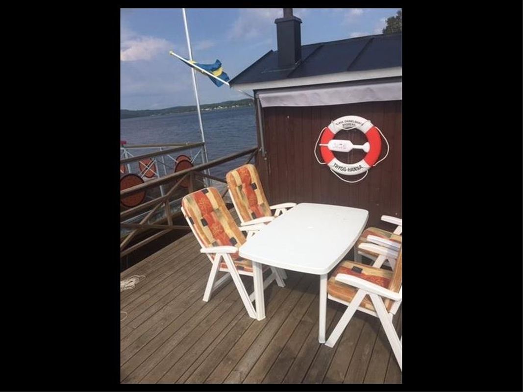 Ombord på flotten, ett vitt bord, fyra vita stolar med gula mönstrade dynor, en röd trävägg där det hänger en livboj, en sbensk fasadflagga vajar i vinden, sjön i bakgrunden.