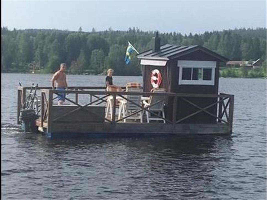 A small red wooden building on a fleet, two persons furniture and a flag.