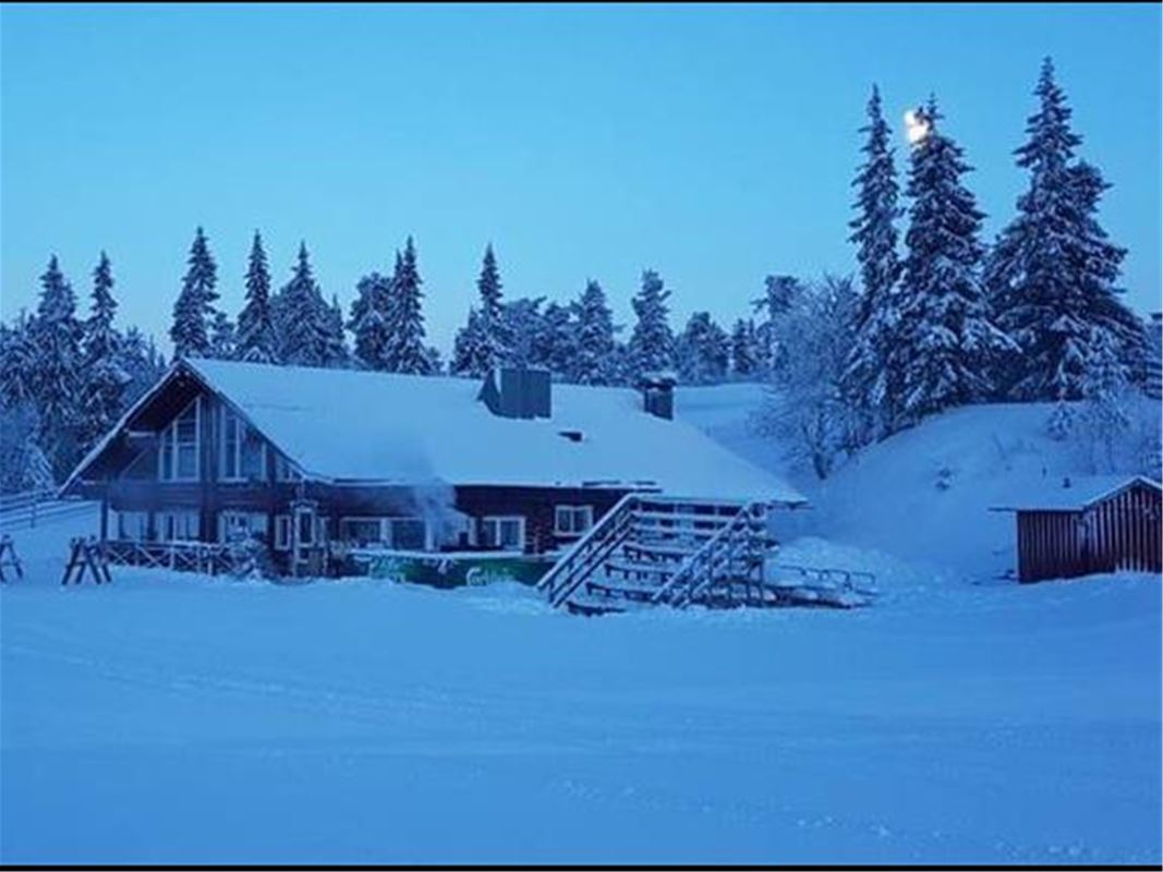 Ett trähus med mycket snö omkring och barrskog bakom huset. 
