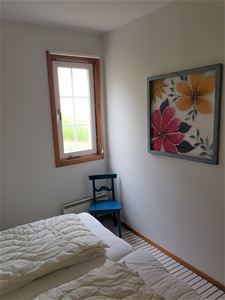 White bedroom with a small window and a painting of flowers on the wall. 