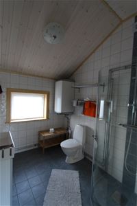 Badrum med vitt kakel och grått klinkers, wc, dusch med glasdörr, stort fönster. 