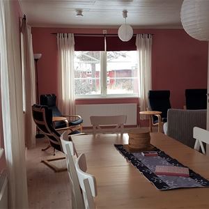 Matbord med träskiva och vita stolar vid ett fönster. I bakgrunden svarta fåtöljer vid ett annat fönster. 