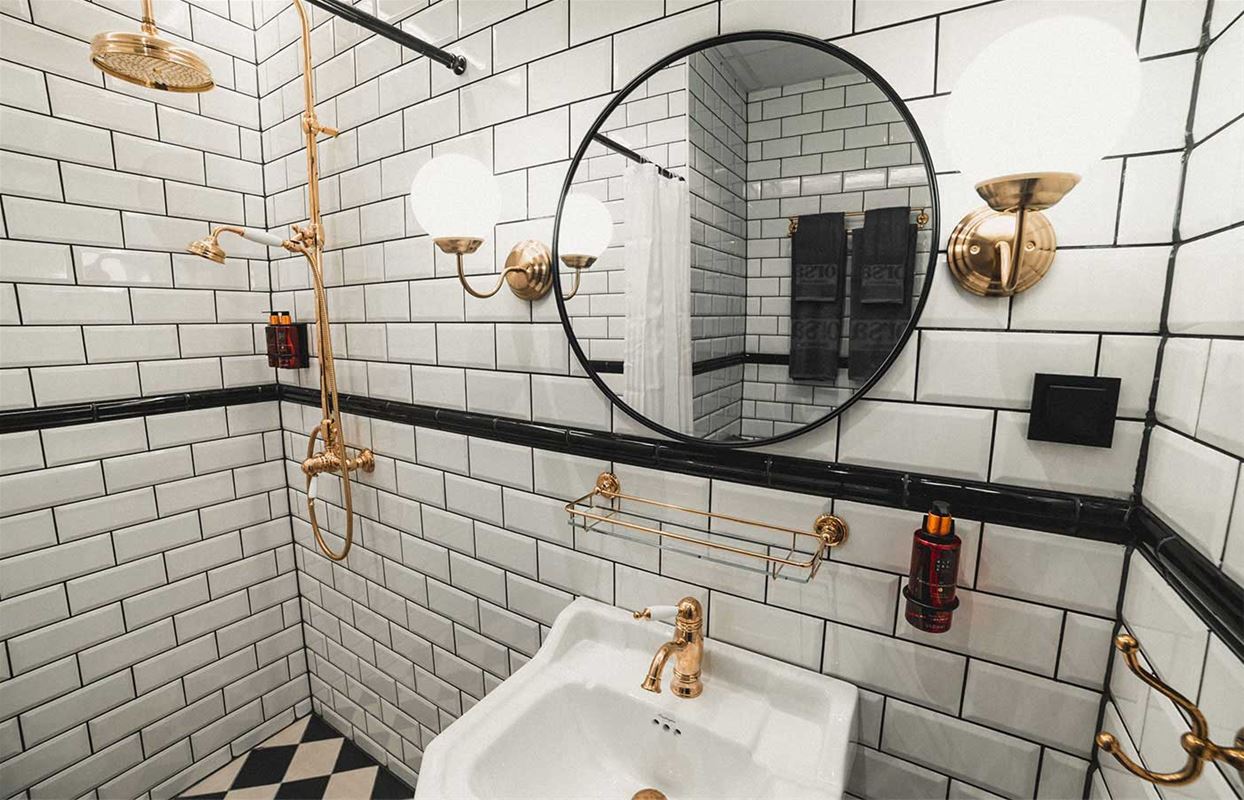Badrum med kaklade, vita väggar och schackrutigt kaklat golv, mässingsfärgade detaljer och en svart, rund spegel. 