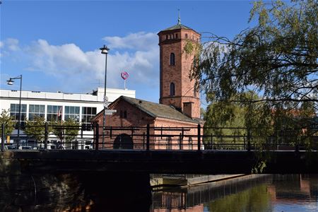 Exterior image, water, bridge with iron railing and a red brick building with a tower.
