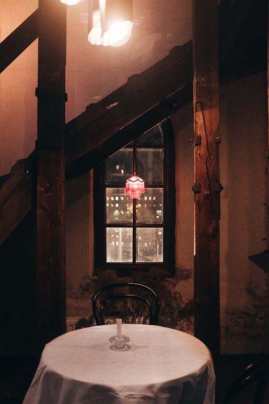 Interiörbild, ett runt litet bord med en vit duk, en ljusstake med ett lus på bordet, ett fönster i bakgrunden.