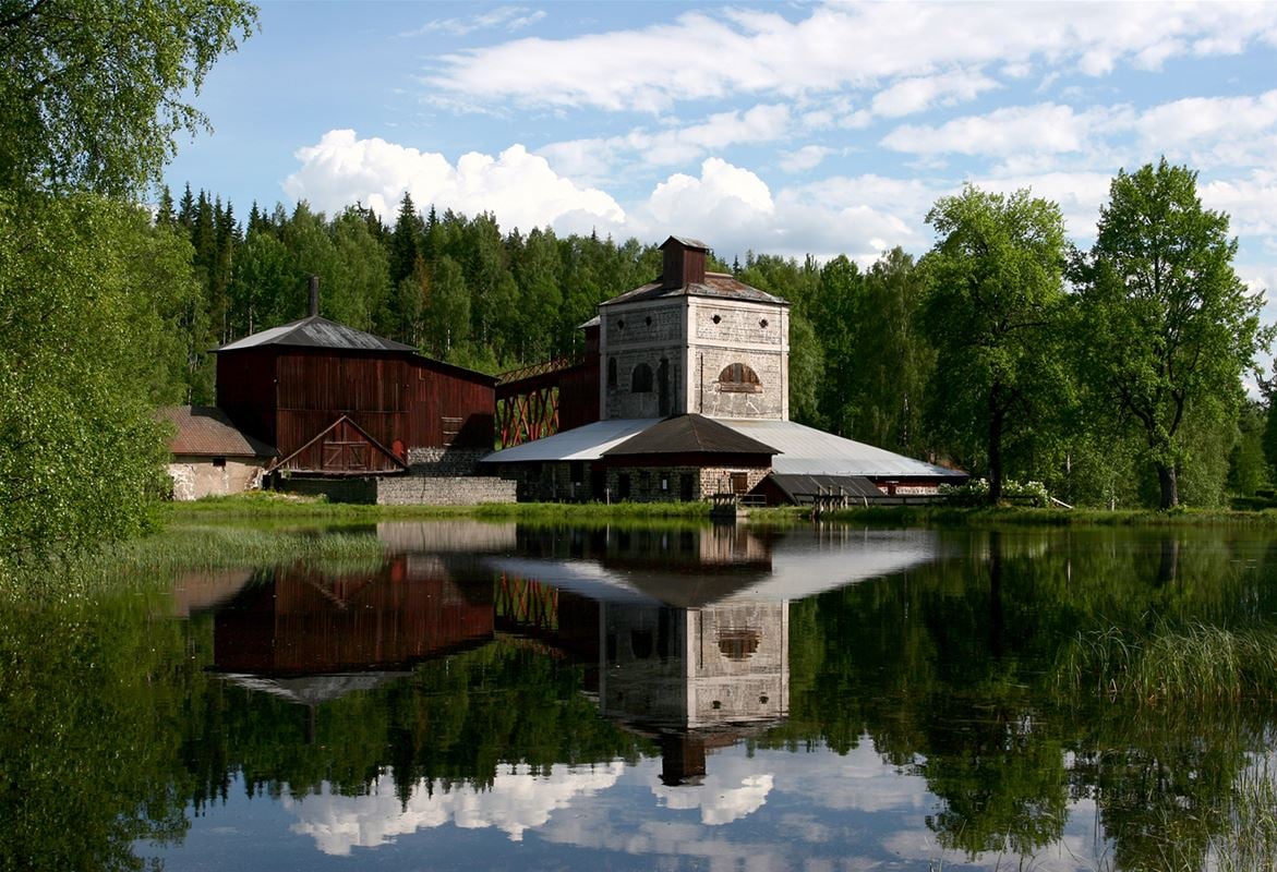 Spegelblankt vatten i förgrund, hyttan vid Ågs bruk, rostugn till vänster och masugn till höger.