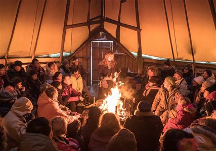 Folk sitter i ring rundt et bål inne i et stort telt med tresøyler.