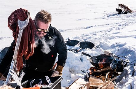 A man holds a shovel in a dug snow pit and the fire burns in an open frying pan.