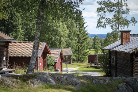Flera röda timmerhus på rad, vatten och blånande berg i bakgrunden.
