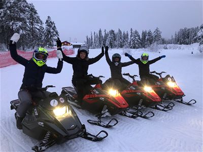 Four snowmobile riders sit on scooters and stretch their arms, the scooters have the light on.