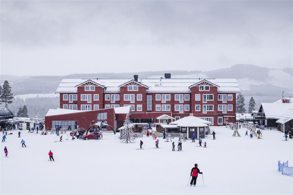 Röd hotellbyggnad i snö med åkande utförsåkare framför.