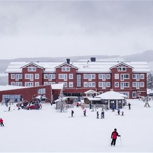 Röd hotellbyggnad i snö med åkande utförsåkare framför.