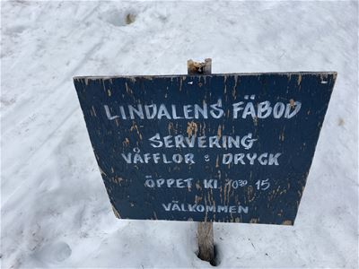 Skylt nedstucken i snön på vilken det står Lindalens fäbod servering med öppettider. 