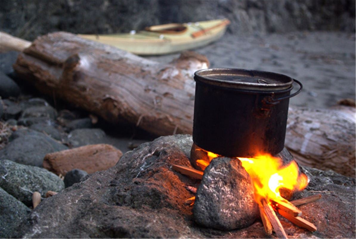 En gryta står på elden ute i skogen. En kajak i bakgrunden. 