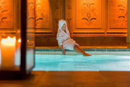 Person in a dressing-gown is sitting on the edge to the indoor pool.