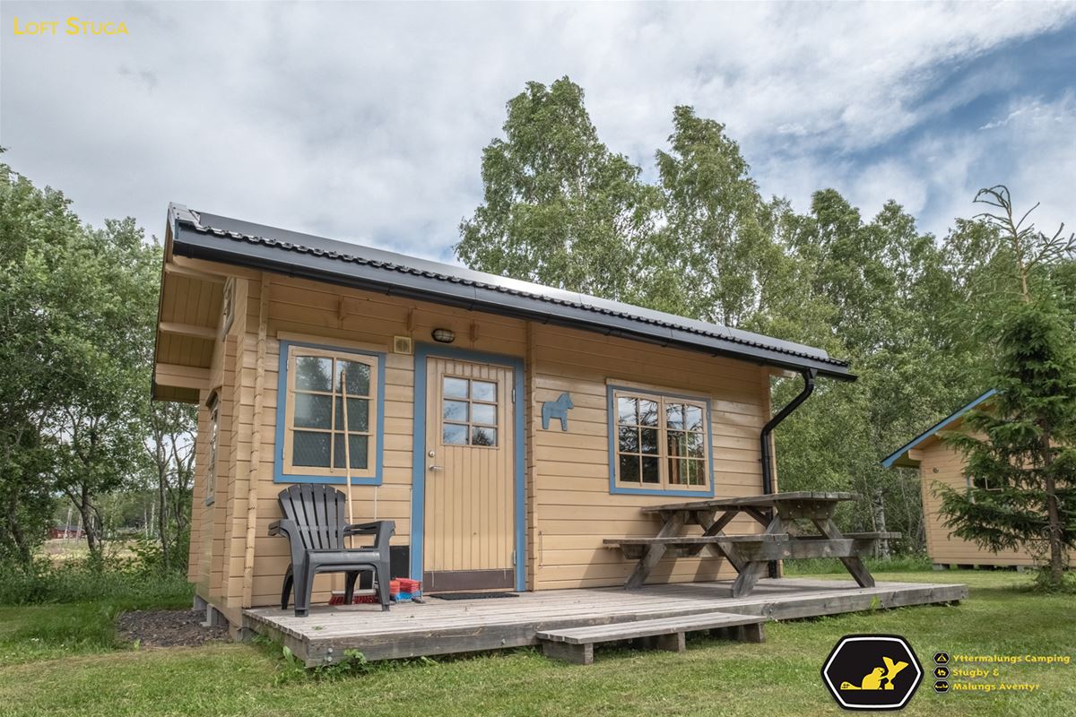 Liten, gul stuga med blåa fönsterfoder och ett picknickbord på altanen.