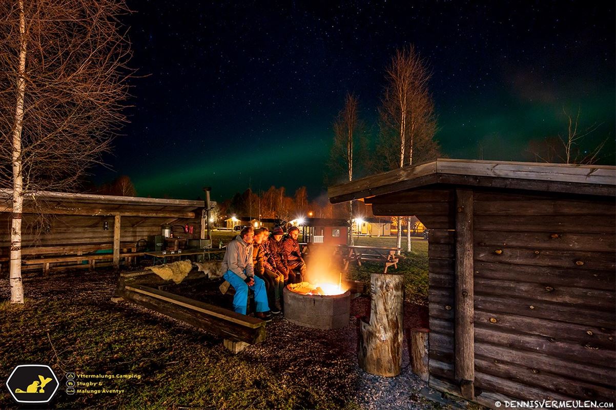 People around a fire in the darkness in front of a small timbered shed.