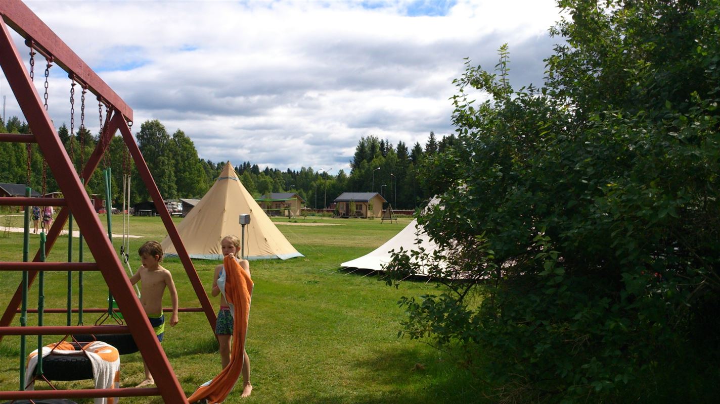 Två barn vid en gungställning och två naturfärgade tält i bakgrunden. 