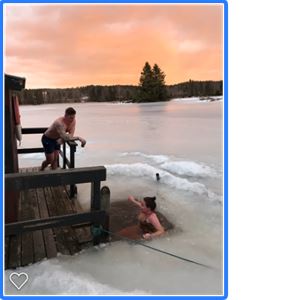 En kvinna badar i en isvak i solnedgången och en man står på en brygga och tittar på. 