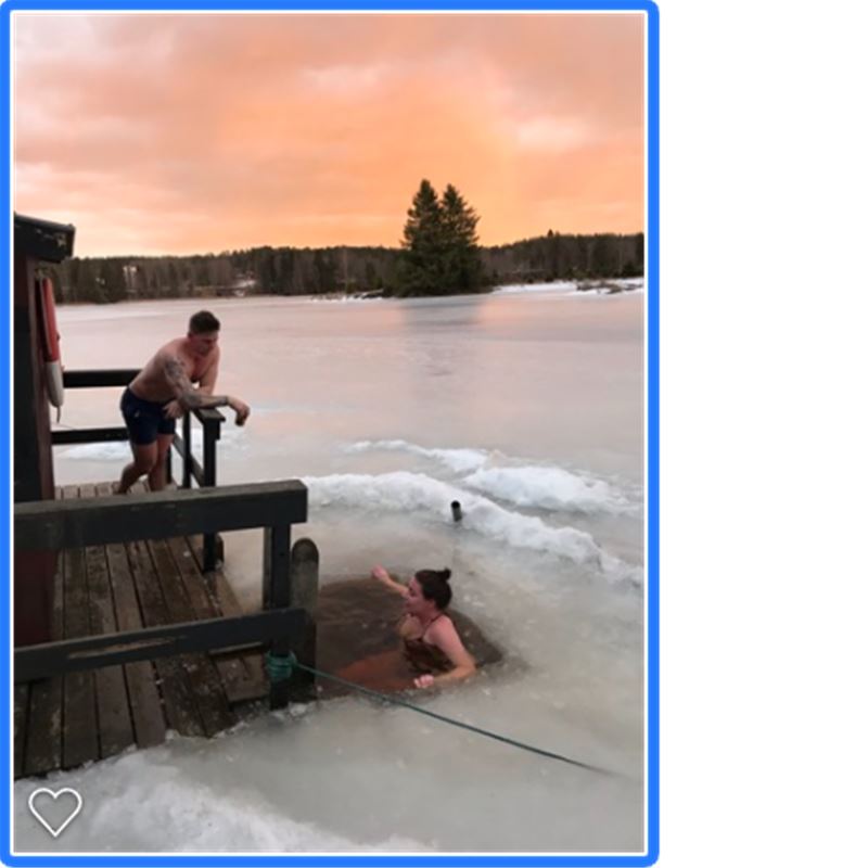 En kvinna badar i en isvak i solnedgången och en man står på en brygga och tittar på. 