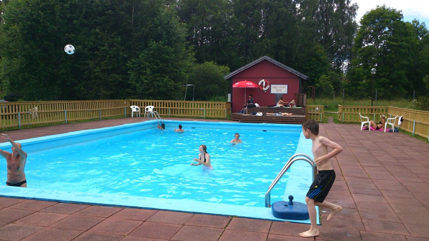 Några personer badar i en utomhuspool omringad av stenplattor och gräsmatta. 