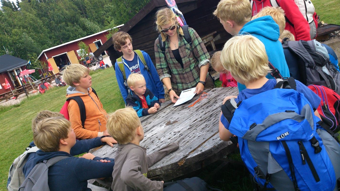 Några barn som står samlade runt ett bord av trä och lyssnar på en ledare. 