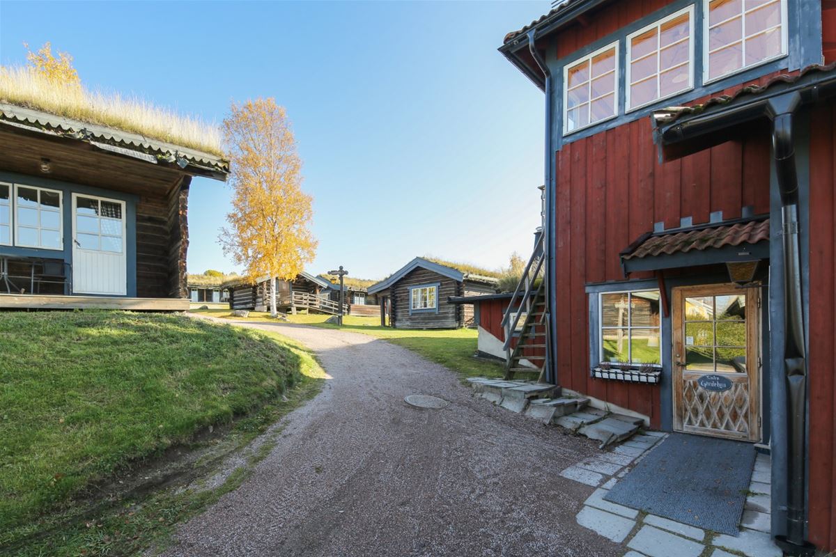 Red house with blue linings and a gravel walk leading to timbered cottages with grass on the roof.