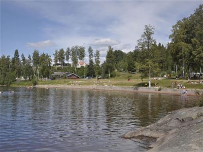 Lake, people at the beach, green area and buildings in the background.