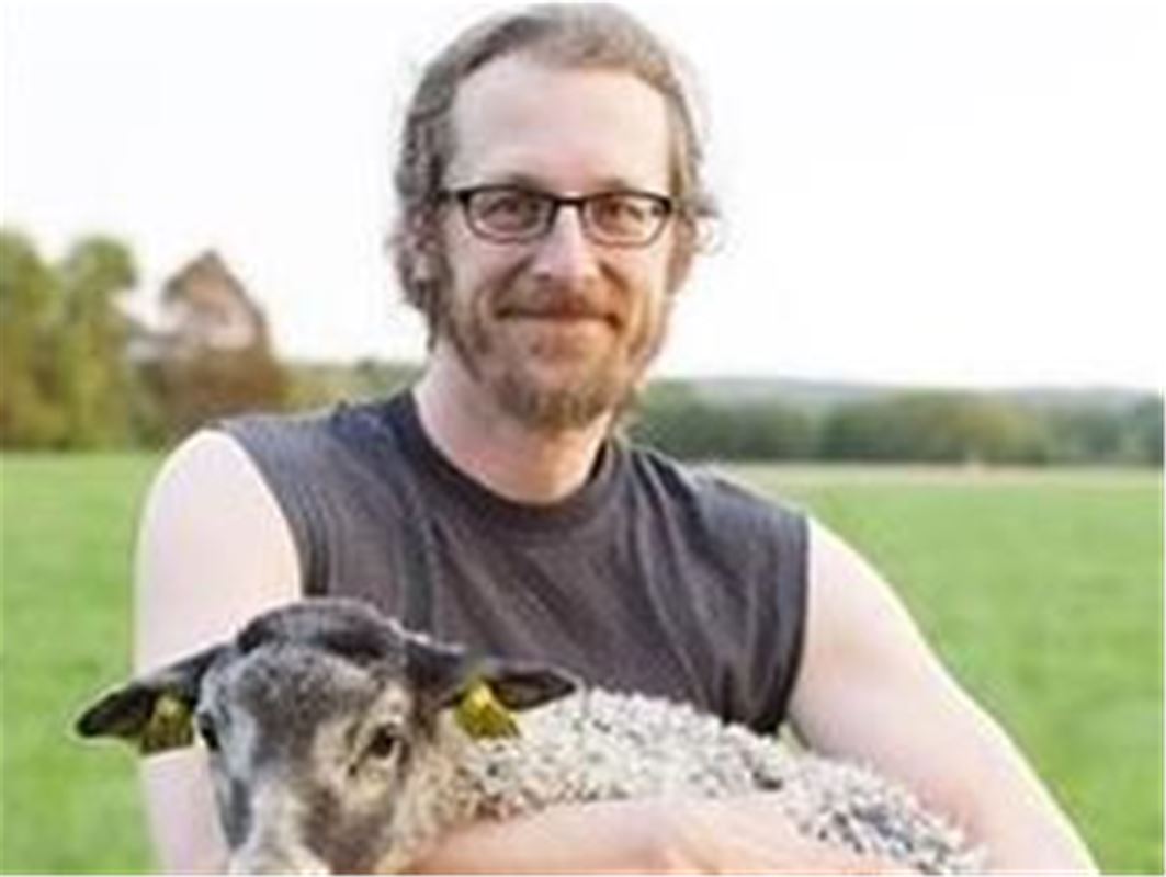 En man i grått linne, skägg och glasögon, mannen håller i ett får.