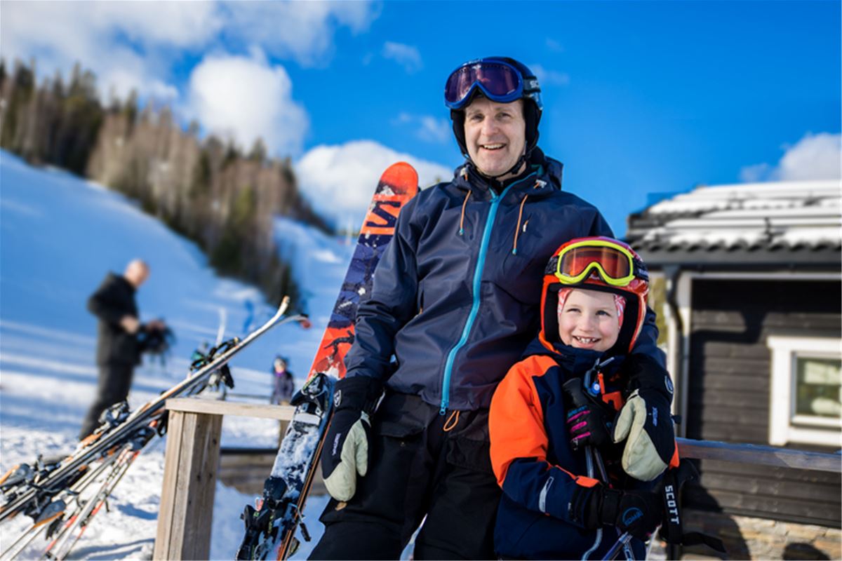 A man and a boy in ski helmets and ski clothes, skiis and a slope in the background.
