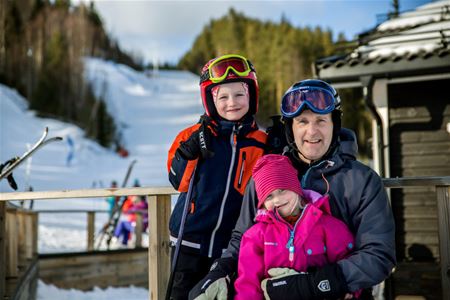 Pappa och två barn, son och dotter i skidkläder, slalombacken och en timmerbyggnad i bakgrunden.