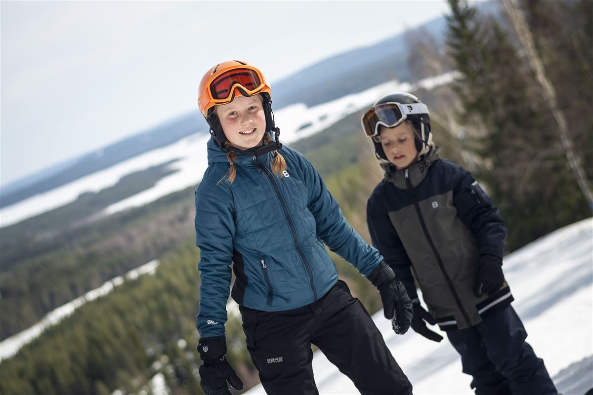 Two children in a ski slope, forest, mountains and a lake in the background.