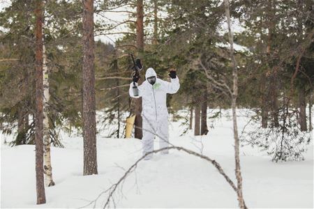 Vitklädd person med paintball gevär uppsträckt med granar omkring i snölandskap.
