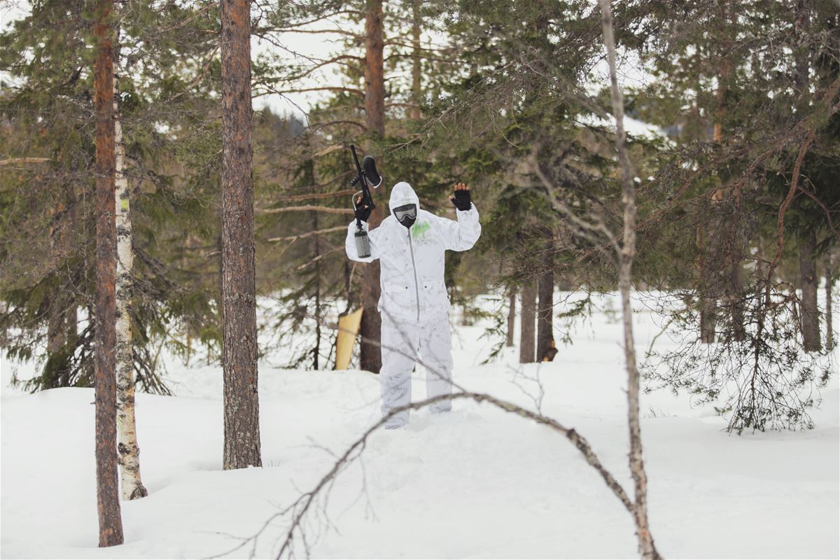 Vitklädd person med paintball gevär uppsträckt med granar omkring i snölandskap.