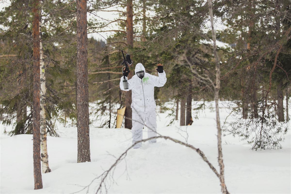 White-clad person with paintball rifle stretched in the air with fir trees around in snowy landscape.