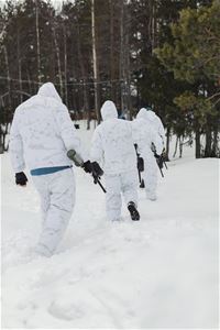 White-clad people walk in a row from the camera in the snow with paitball rifles in their hands.